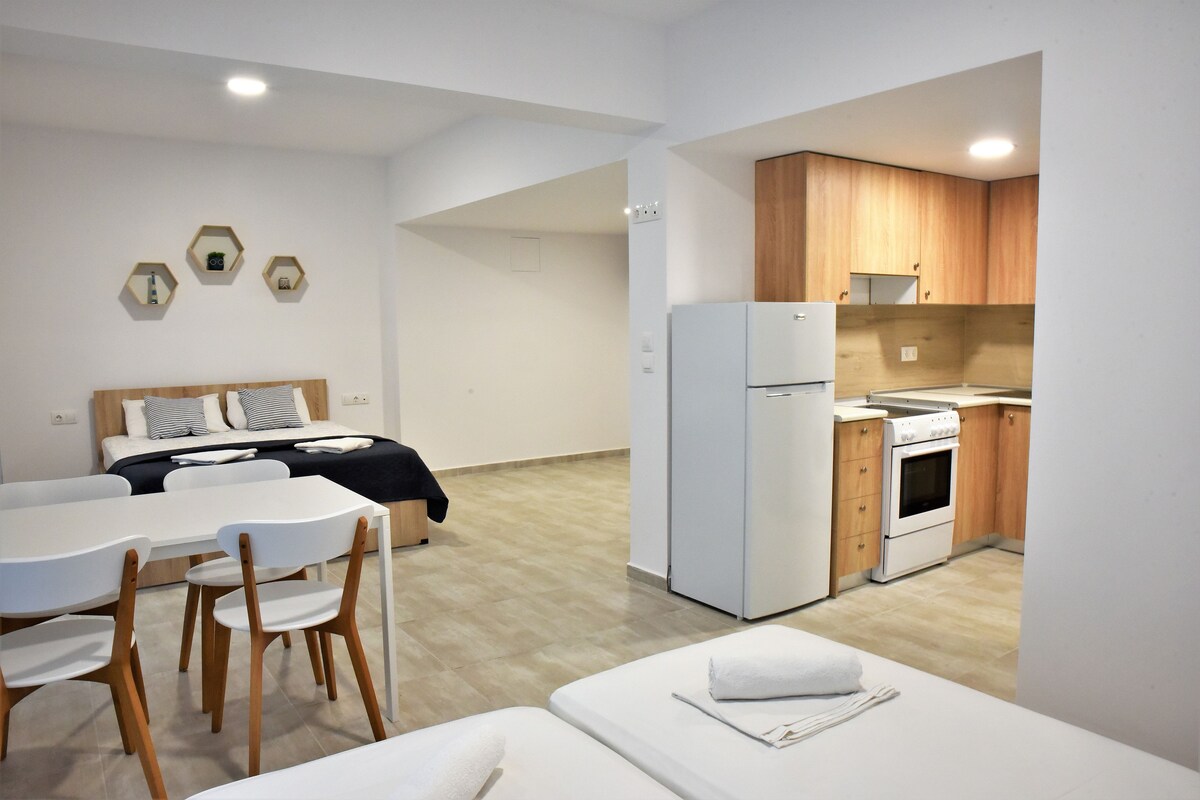 A modern hotel room with a bed, a dining table with chairs, a kitchen area with a refrigerator and stove, and a small seating area with a bench.