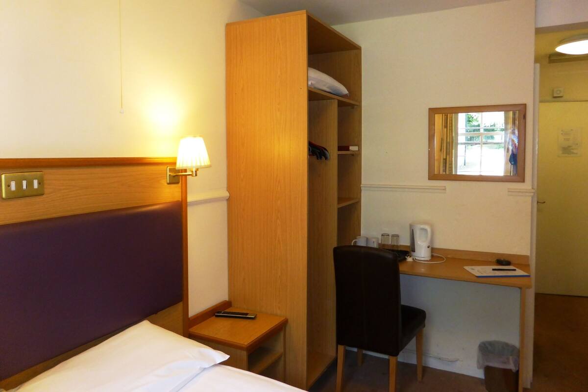 A hotel room with a bed, a desk with a chair, a wooden wardrobe, a nightstand with a lamp, and a window with a view.