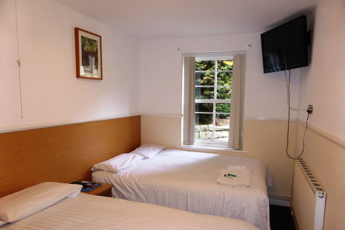 A small, clean room with two single beds, a window with curtains, a flat-screen TV mounted on the wall, and a radiator.