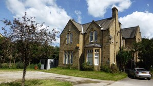 A large stone building with multiple windows and a chimney, surrounded by greenery and a parking area. - The Old Vicarage Hotel (Leeds)