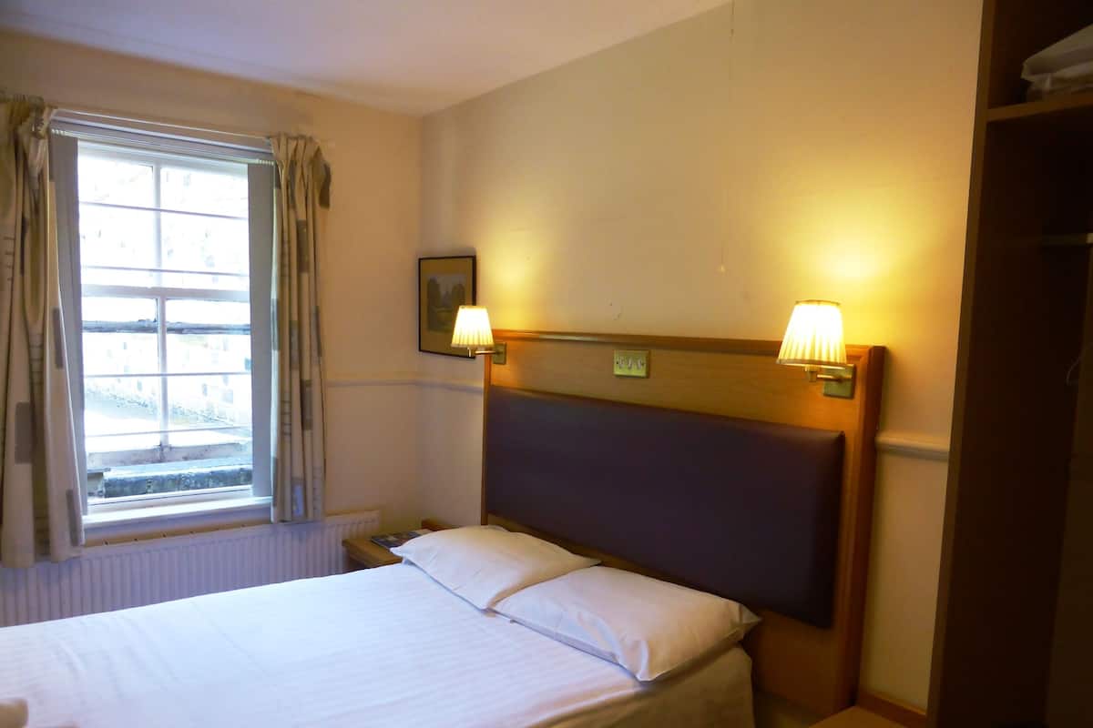 A hotel room with a bed, two bedside lamps, a window with curtains, and a radiator.