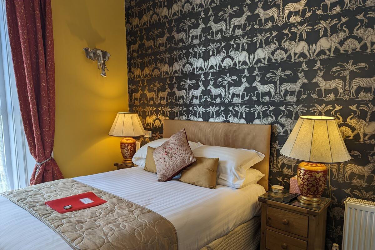 A neatly made bed with a quilt, pillows, and a red folder on the bed. There are two bedside lamps on wooden nightstands. The room has a yellow wall and a patterned wallpaper with animal prints.