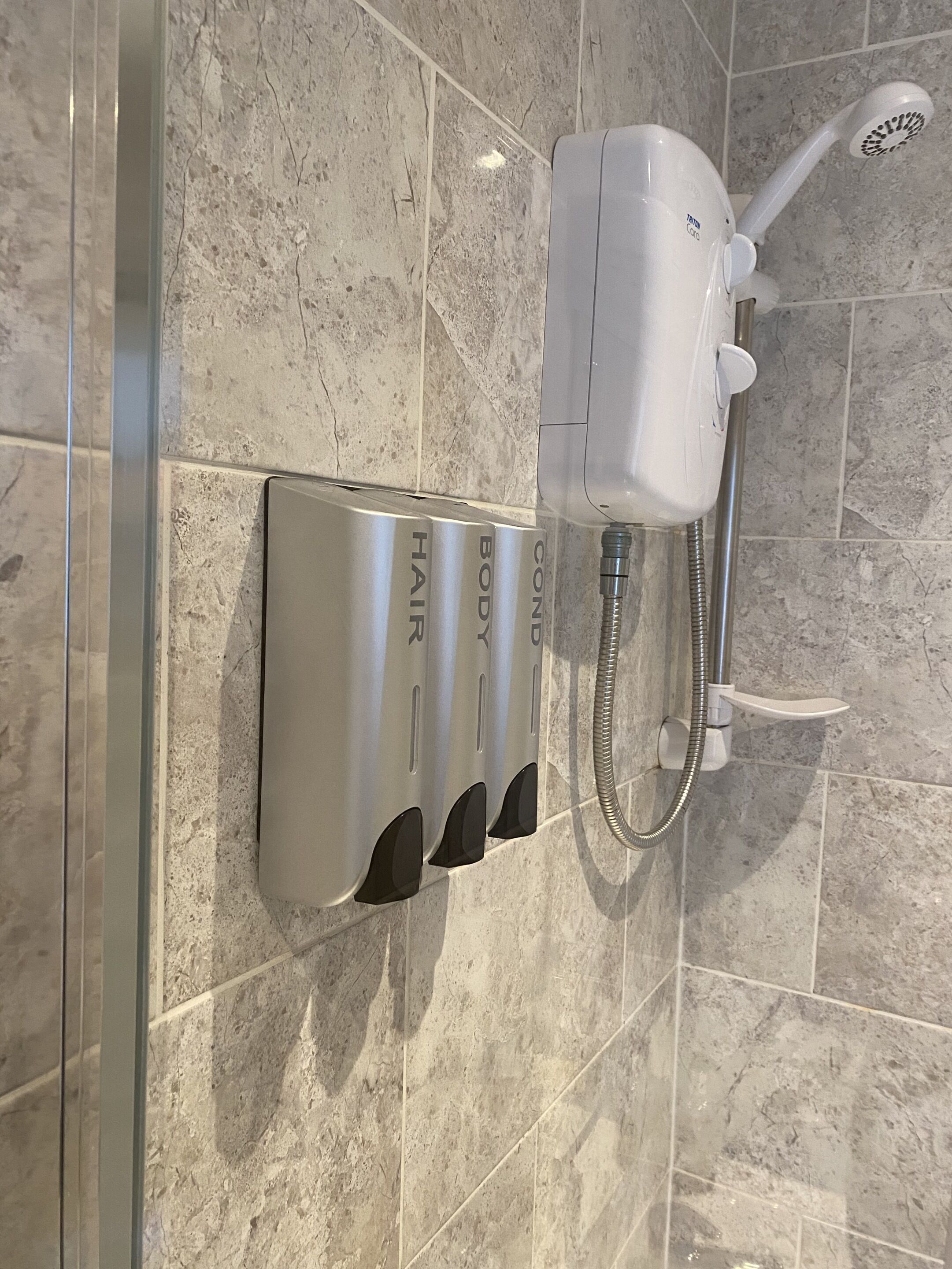 Ein Badezimmer mit Dusche, Duschbrause und drei an der Wand montierten Handtrocknern, beschriftet mit 'HAIR', 'BODY' und 'CONDITIONER'.