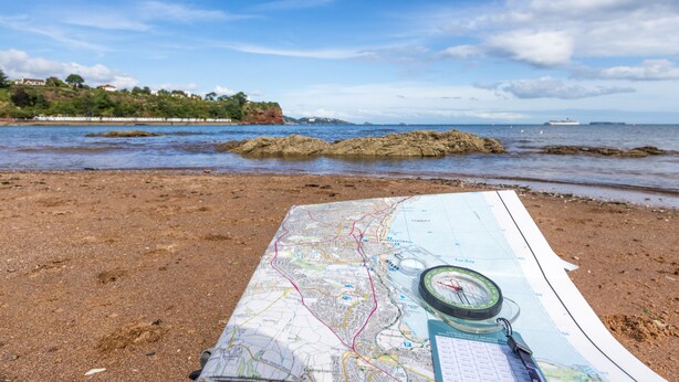Une carte et une boussole sur une plage de sable, avec un affleurement rocheux et un navire au loin.