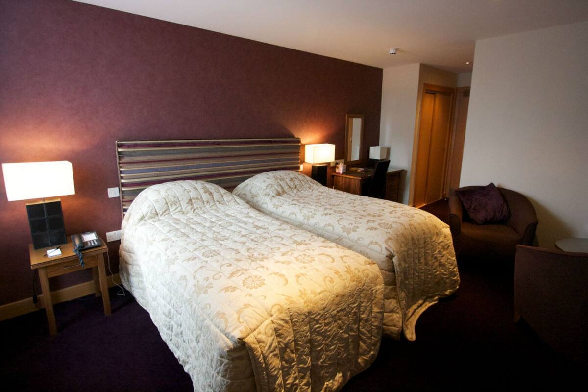 A hotel room with two beds, a desk with a lamp, a chair, and a door.