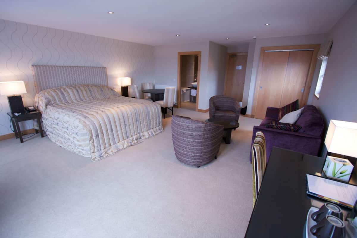 A hotel room with a large bed, a sitting area with a table and chairs, a small sofa, and a desk with a lamp and a tray of glasses.