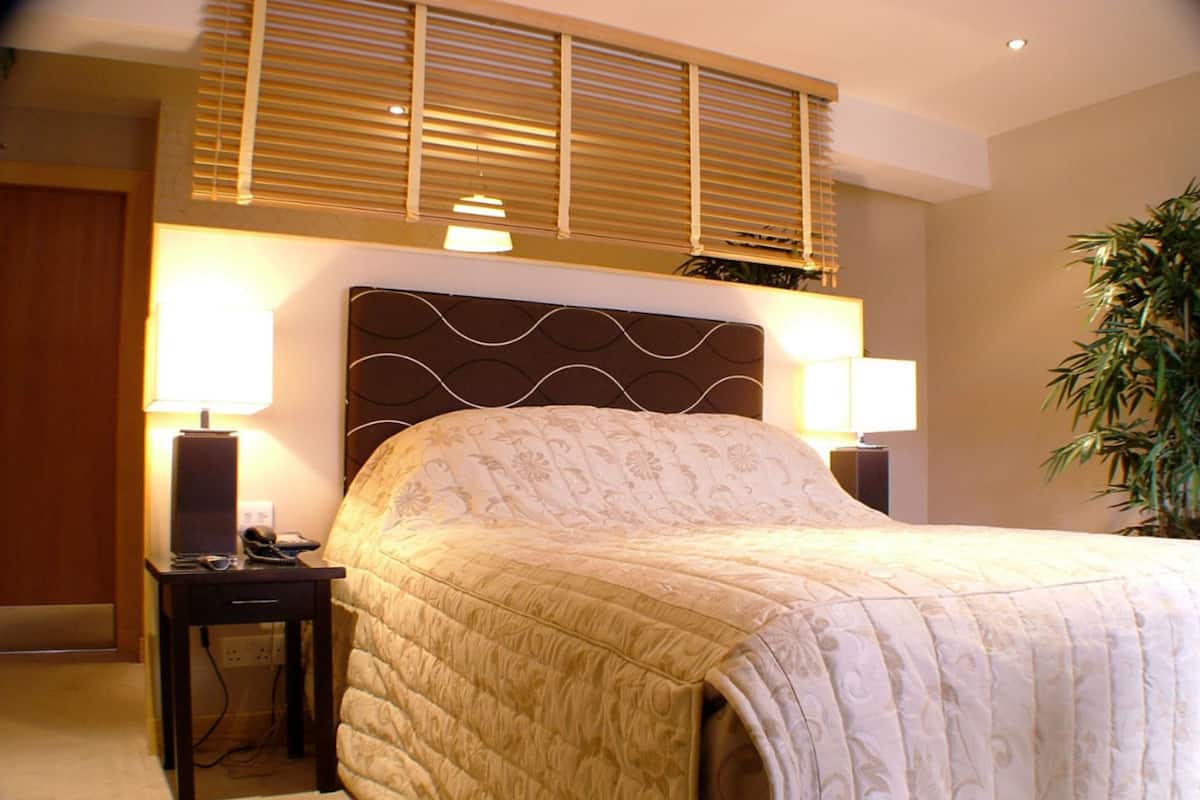 A hotel room with a large bed, two bedside lamps, a nightstand, a wall-mounted shelf, and a potted plant.