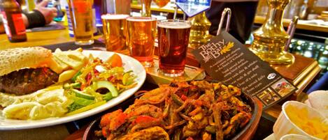 Un bar con varios platos, incluyendo una fuente de carne a la parrilla, una ensalada y un bol de sopa. Se ven varias llaves de cerveza y vasos.