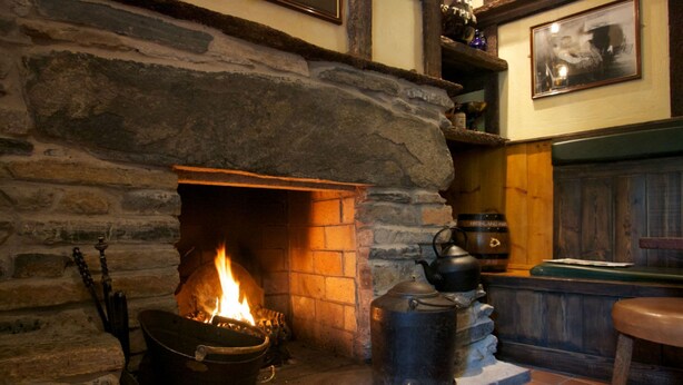 A stone fireplace with a fire, a kettle, and a wooden shelf with bottles.