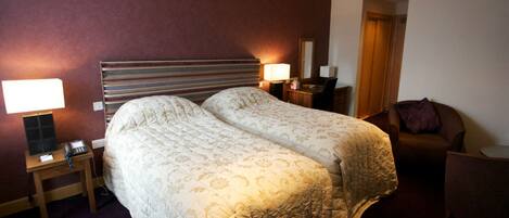 Una habitación de hotel con una cama doble, mesitas de noche con lámparas, una silla y una puerta que da a otra habitación.