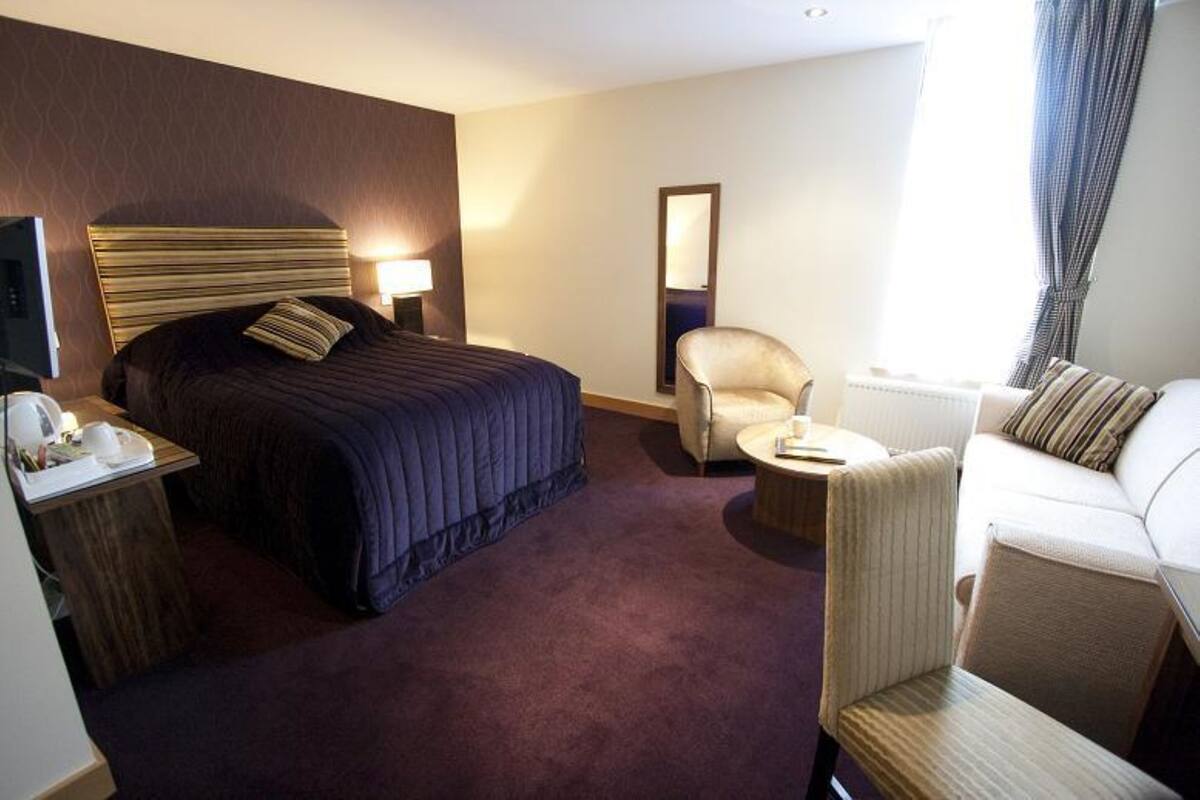 A hotel room with a bed, a desk with a lamp, a chair, a sofa, and a large mirror.