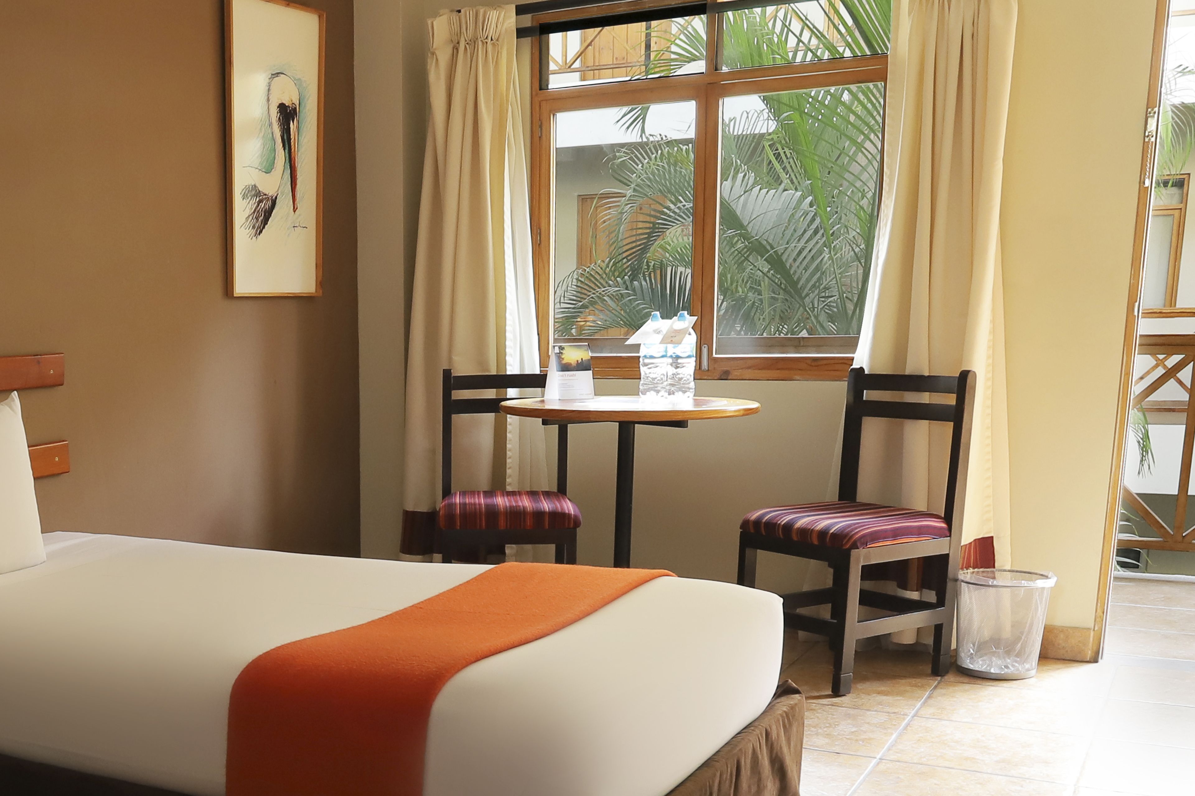 Une chambre d’hôtel avec un lit, deux chaises, une petite table, un tableau encadré et une fenêtre avec des rideaux.