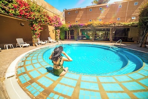 A pool with a tiled edge, a woman sitting by the pool, and a building with a balcony in the background.