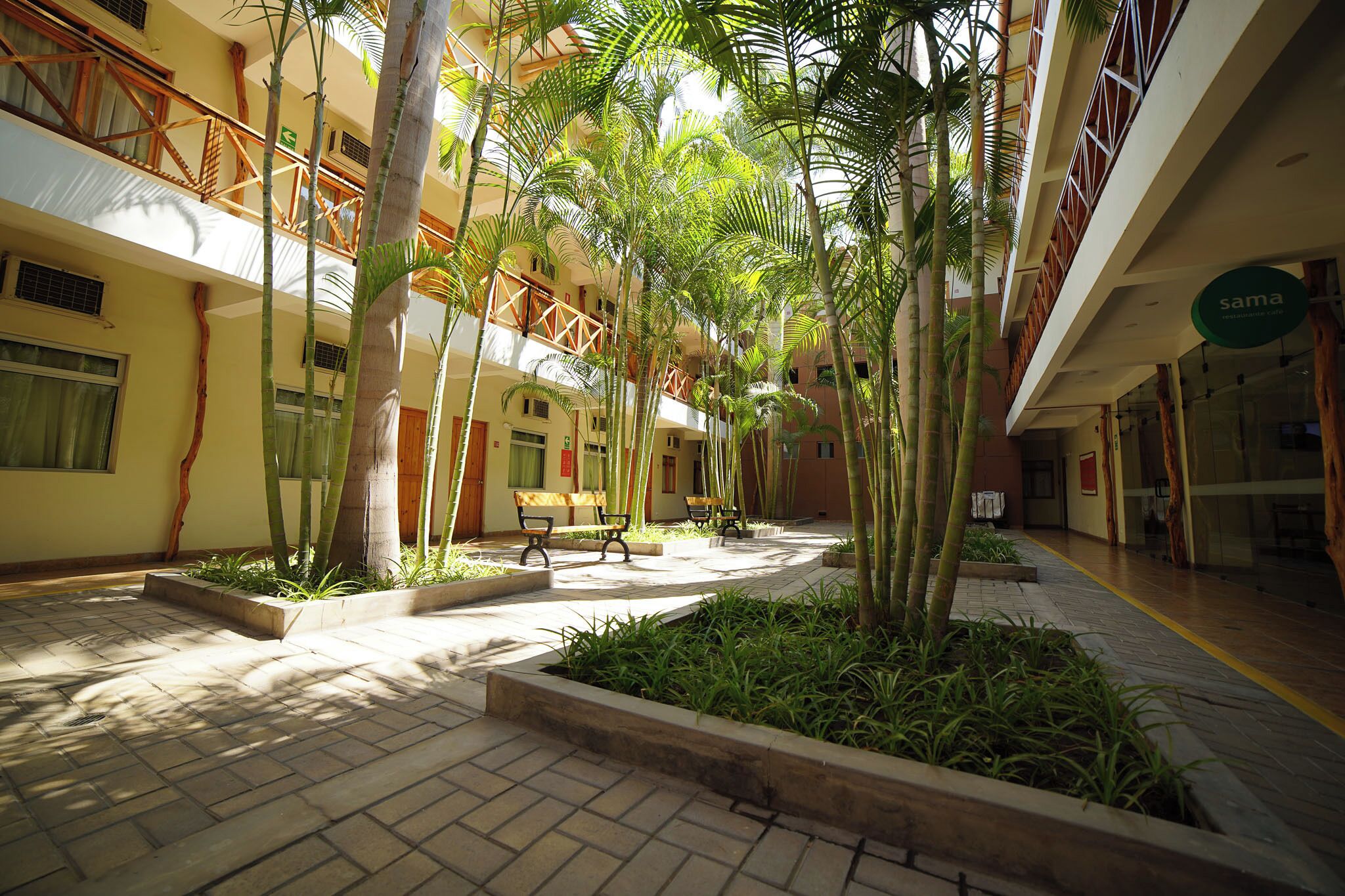 Un patio con jardín central, bancos y un edificio con el rótulo 'sama'.