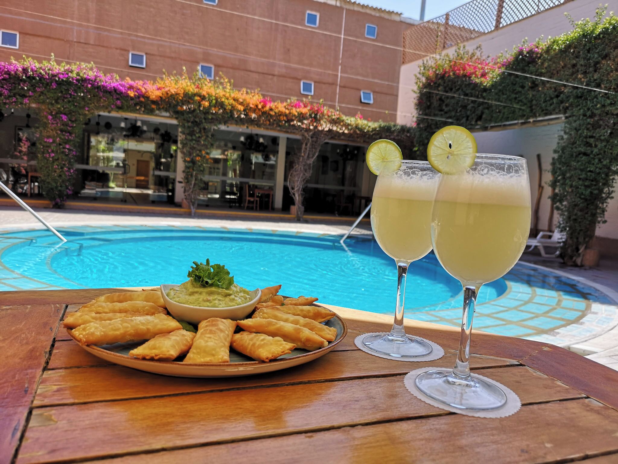 A poolside table with a plate of samosas, a bowl of dip, and two glasses of a yellow beverage, likely lemonade, with lemon slices.
