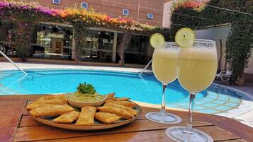 Mesa junto a la piscina con un plato de samosas, un bol de salsa y dos vasos de una bebida amarilla, probablemente limonada, con rodajas de limón.