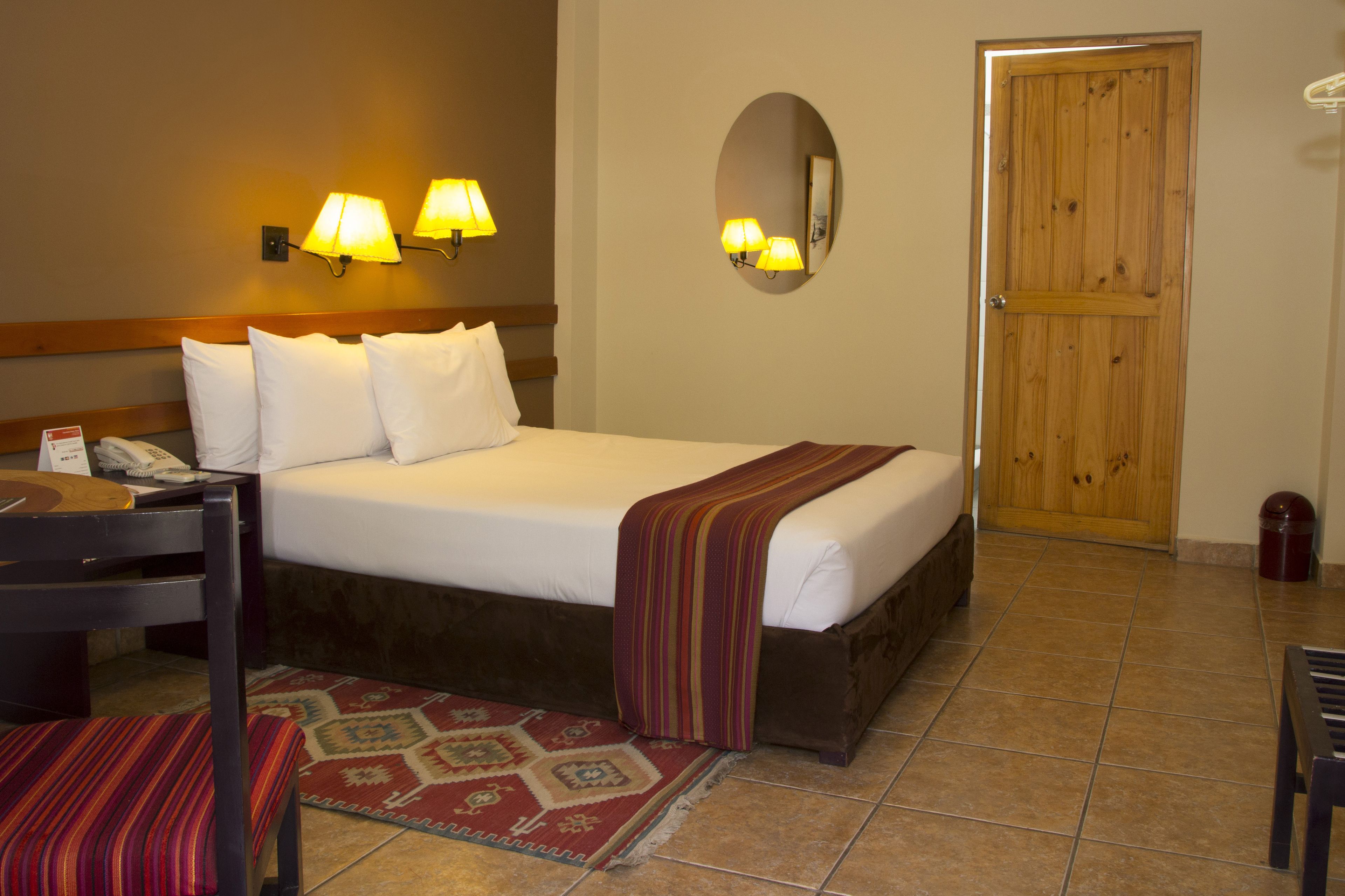 A hotel room with a bed, bedside lamps, a telephone, a chair, a rug, and a wooden door.