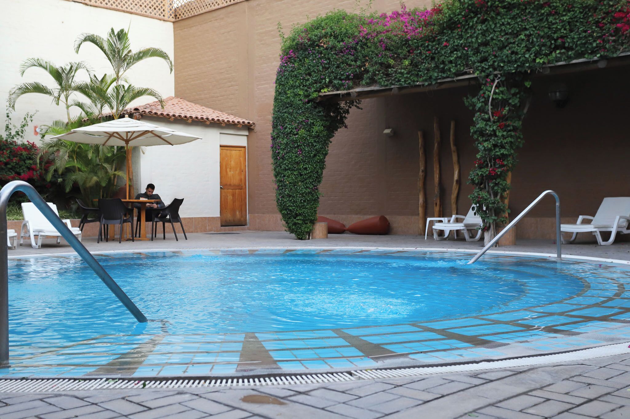 Ein Pool mit Liegestühlen und einem Tisch, umgeben von Pflanzen und einer gefliesten Terrasse.
