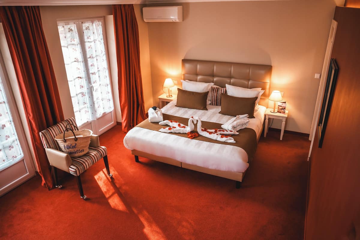 A hotel room with a large bed, two bedside tables with lamps, a chair with a basket, and a window with curtains.