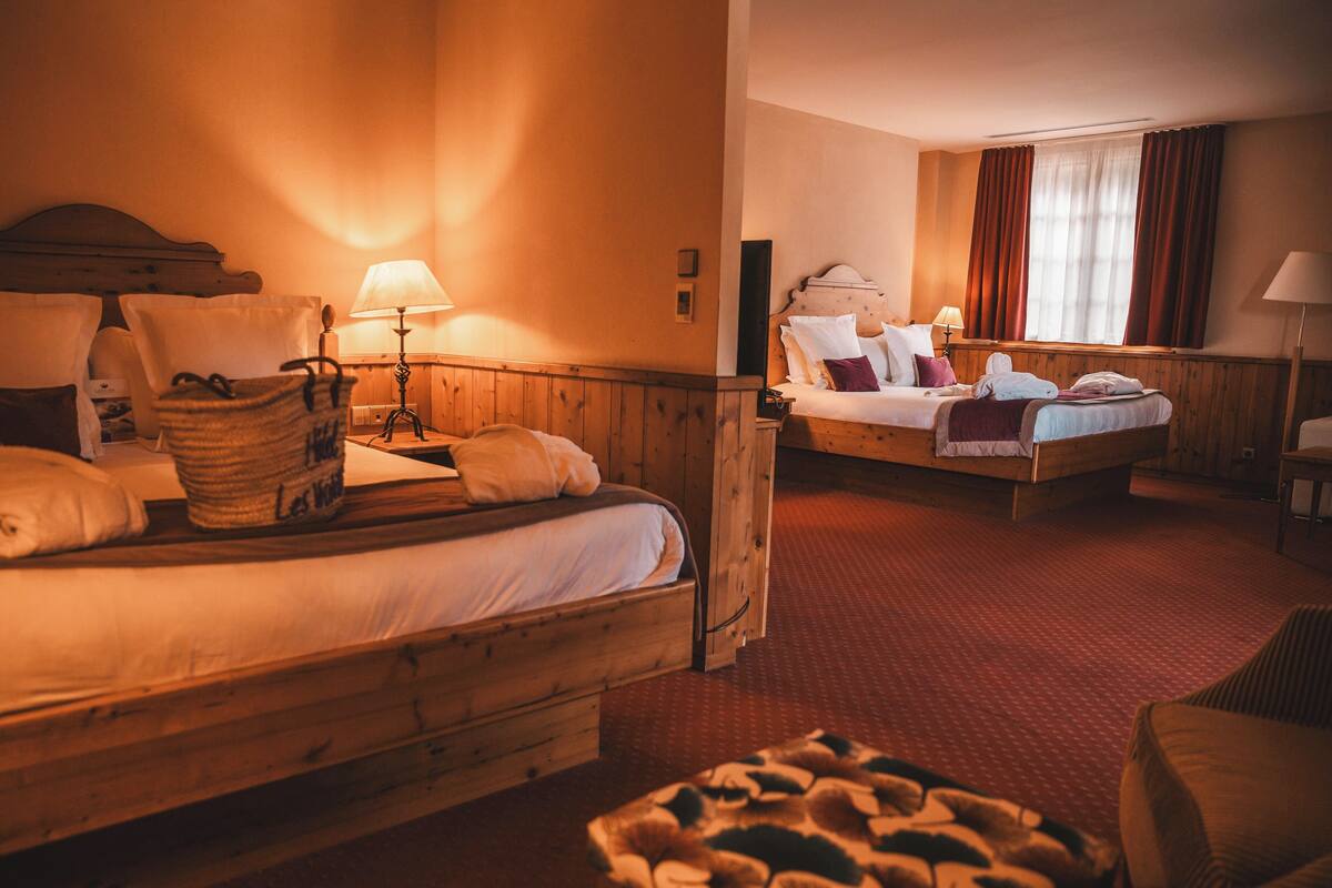 A hotel room with two beds, wooden headboards, and a red carpet. There are lamps on each bed and a basket on the left bed.