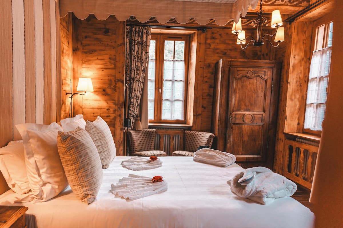 A wooden cabin-style room with a bed, pillows, a chair, a lamp, and a window.