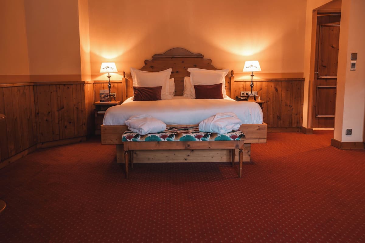 A neatly made bed with pillows and a blanket, wooden headboard, two bedside tables with lamps, and a red carpeted floor.