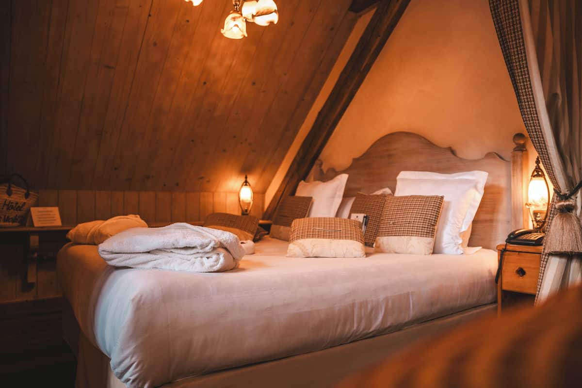 A cozy, wooden-floored bedroom with a large bed, pillows, and a nightstand with a lamp.