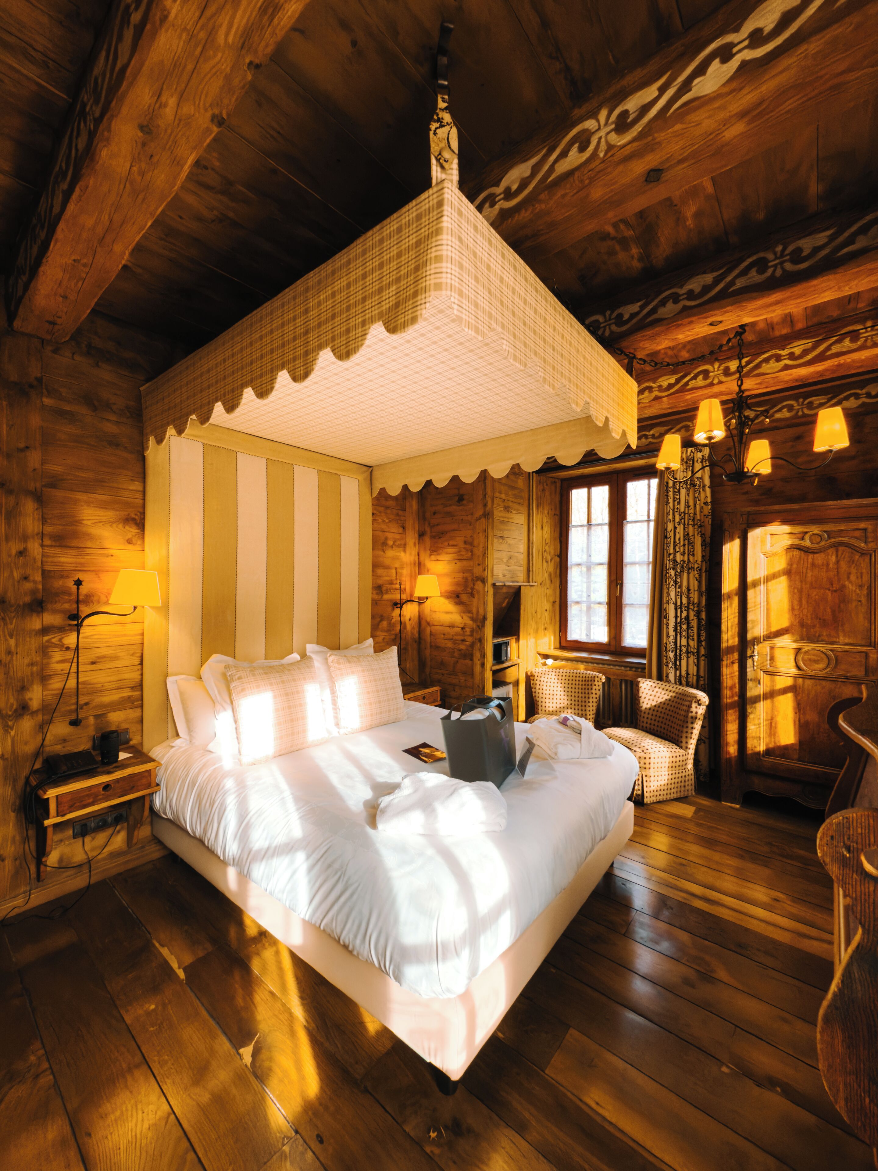 A wooden cabin bedroom with a four-poster bed, a nightstand, a chair, and a hanging light fixture.