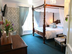 A four-poster bed with a canopy, a wooden desk with a vase of flowers, and a green kettle on a side table. - The Arches B&B (St Austell)