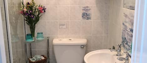 A bathroom with a toilet, sink, and shower. There is a vase with flowers on a shelf.