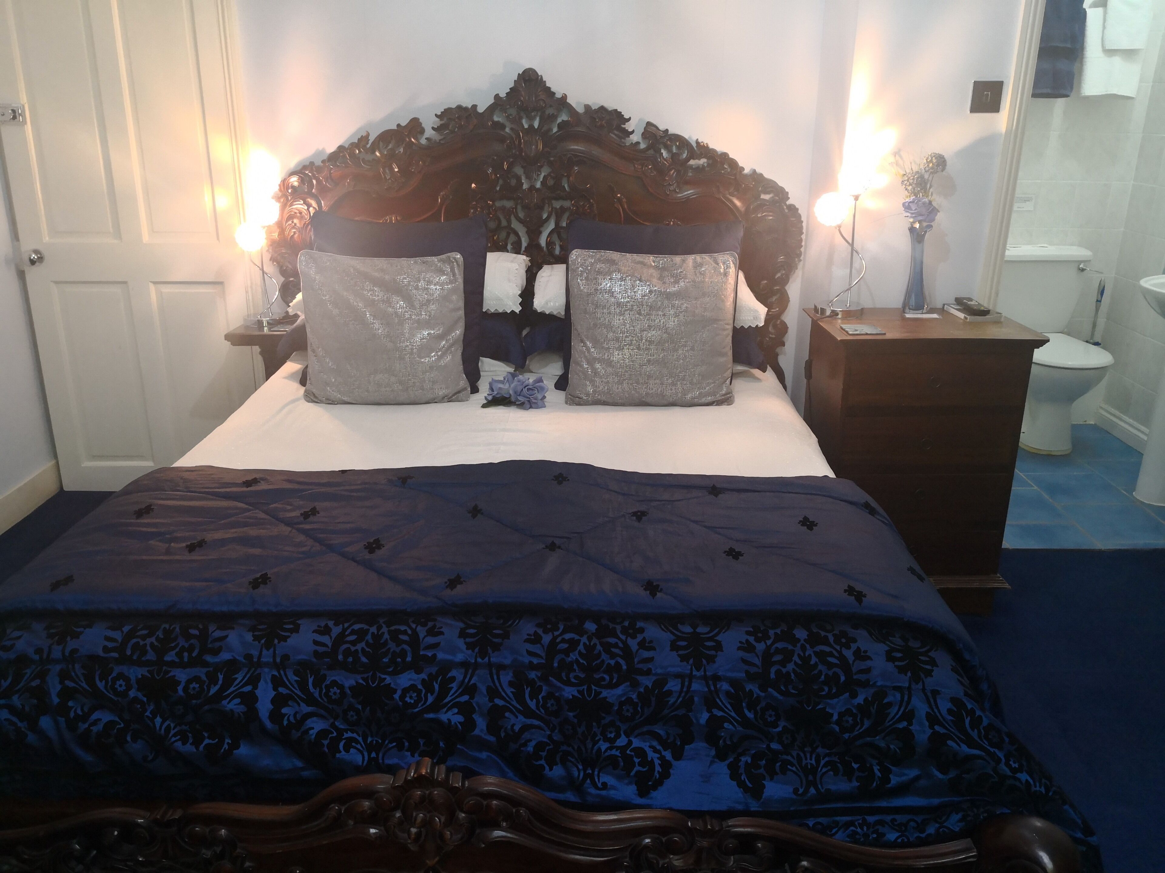 Een net opgemaakt bed met een donkerblauwe sprei en twee grote kussens, een houten hoofdbord, een nachtkastje met een lamp, en een badkamer op de achtergrond.