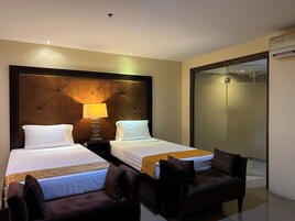 A hotel room with two beds, a nightstand with a lamp, a seating area with chairs, and a sliding door leading to a shower area.