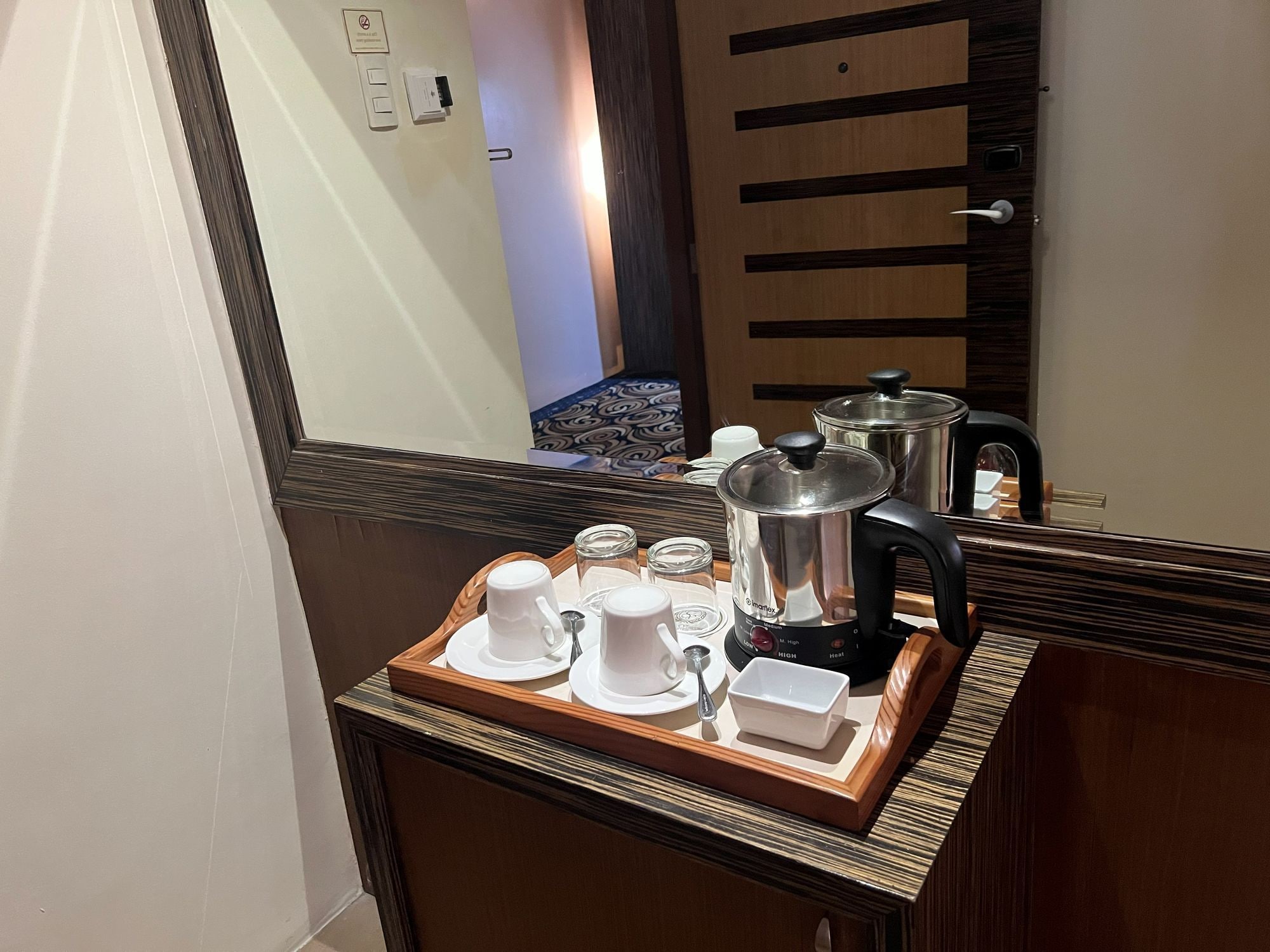 A hotel room with a wooden sideboard, a tray with white cups and a kettle, and a large mirror reflecting the room.