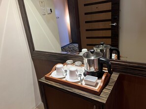 A hotel room with a wooden sideboard, a tray with white cups and a kettle, and a large mirror reflecting the room.