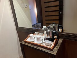 A hotel room with a wooden sideboard, a tray with white cups and a kettle, and a large mirror reflecting the room.