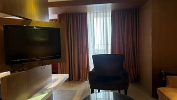 Een hotelkamer met een flatscreen televisie bevestigd aan een houten paneel, een zwarte fauteuil, een ronde salontafel met een afstandsbediening, en een raam met gordijnen.
