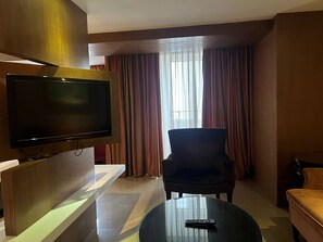 A hotel room with a flat-screen TV mounted on a wooden panel, a black armchair, a round coffee table with a remote control, and a window with curtains.