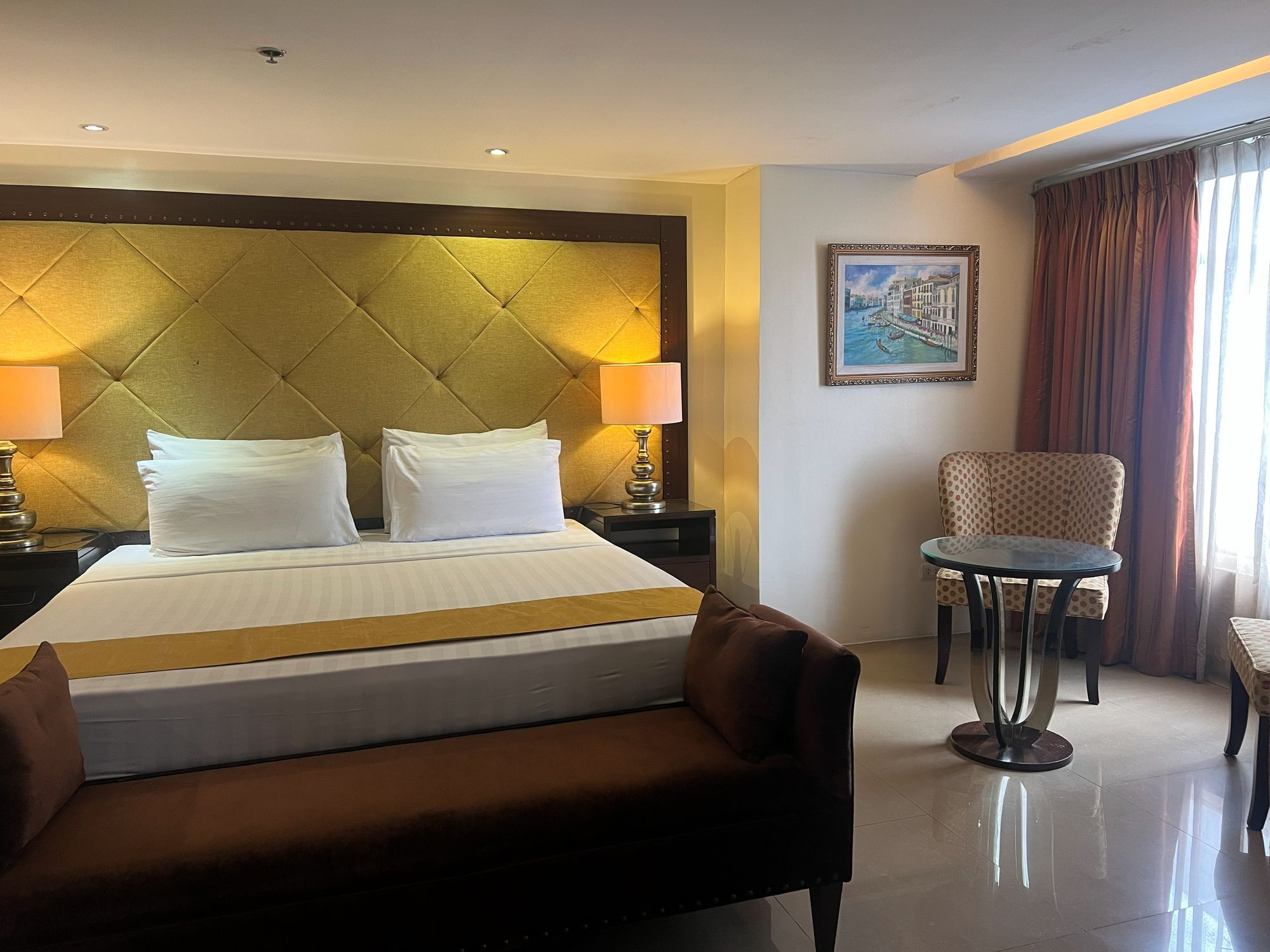 A hotel room with a large bed, two bedside lamps, a chair, a small table, and a painting on the wall.