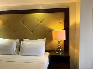 A hotel room with a tufted headboard, a bed with white linens, two pillows, a bedside table with a lamp, and a nightstand.
