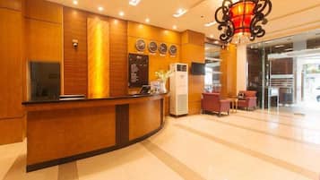 Zona de recepción de hotel con un mostrador de madera, un reloj digital y un área de descanso con sillones tapizados en rojo.