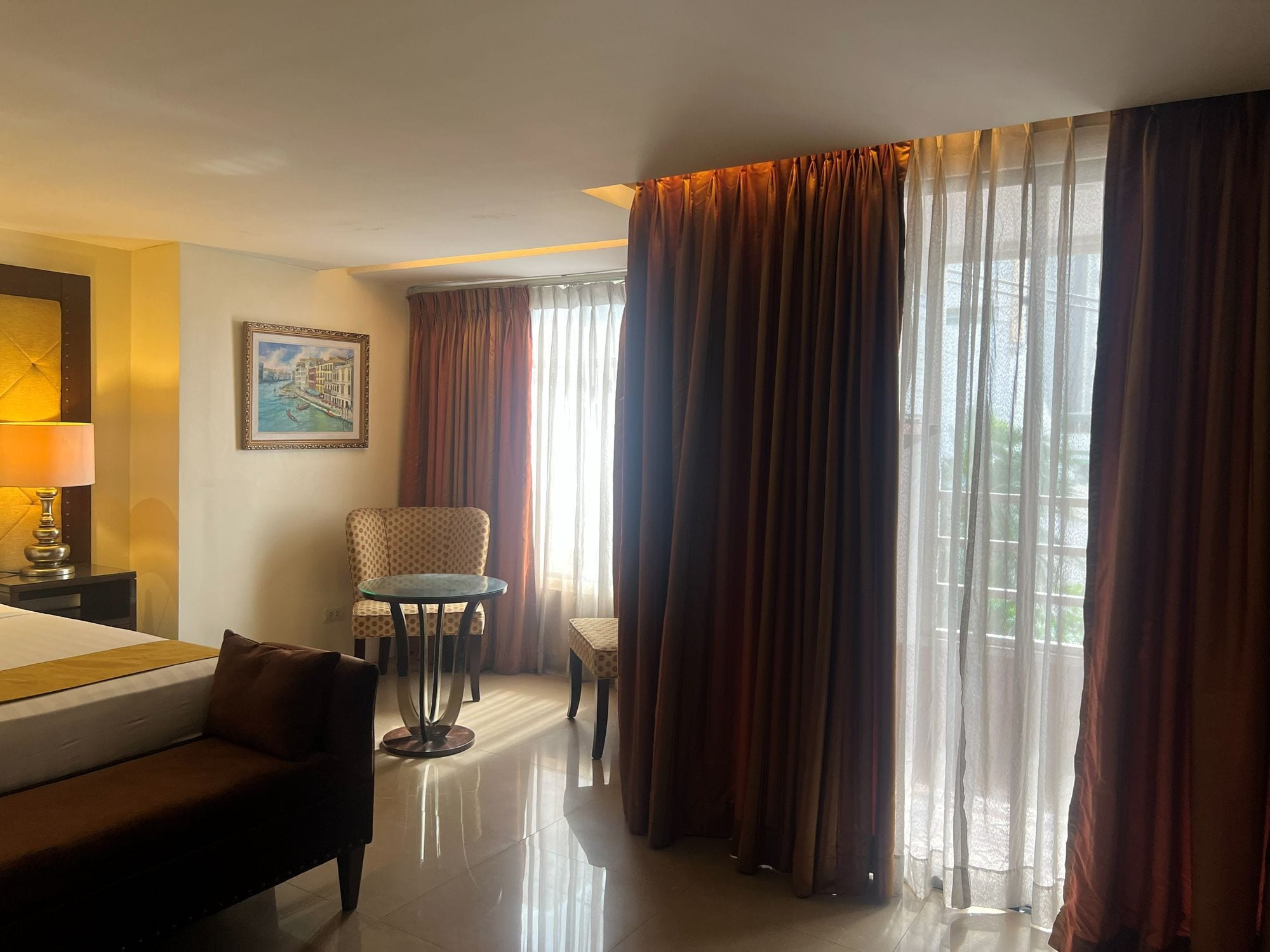 A hotel room with a bed, a chair, a small table, and a painting on the wall.