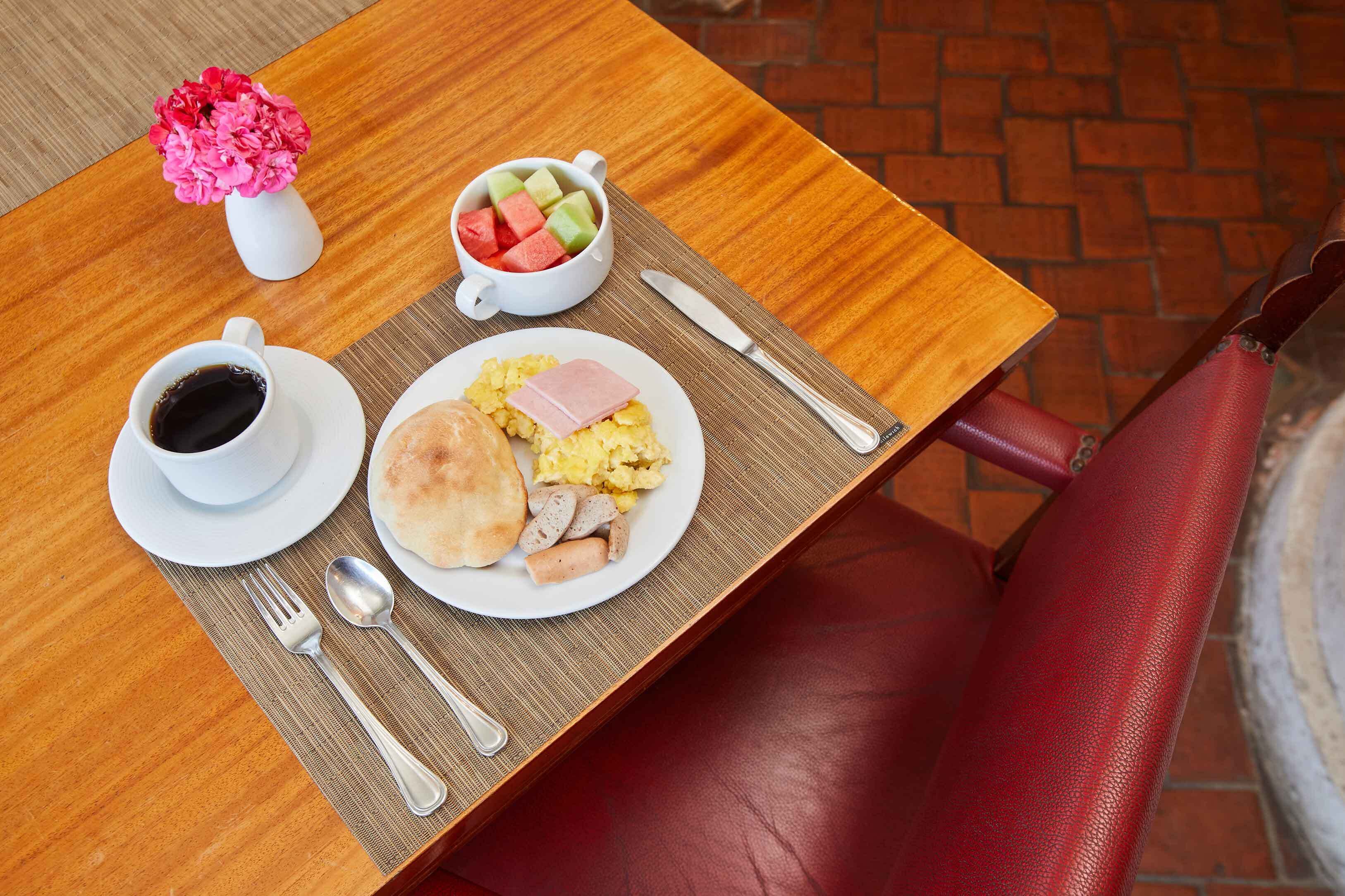 Una mesa de desayuno con una taza de café, un plato de huevos, salchicha y un bol de fruta.
