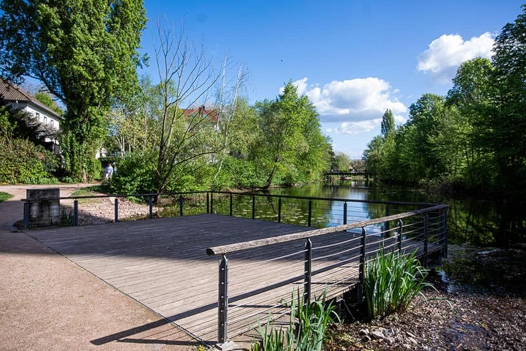 A wooden bridge over a calm river, surrounded by lush greenery and trees.