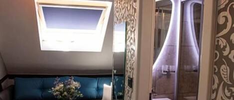Un espacio habitable pequeño con un sofá, una mesita redonda, un televisor montado en la pared y un baño visible a través de una puerta abierta.