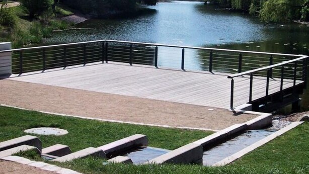 A wooden deck with a metal railing overlooking a river, surrounded by greenery and a stone pathway. - Hotel Rosengarten Kehl (Kehl)