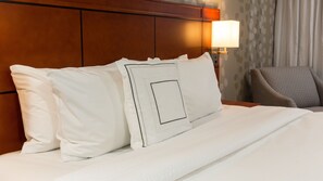 Un lit bien fait, avec du linge de lit blanc et des oreillers, une tête de lit en bois, une lampe de chevet et une chaise agrémentée d’un coussin à motifs.
