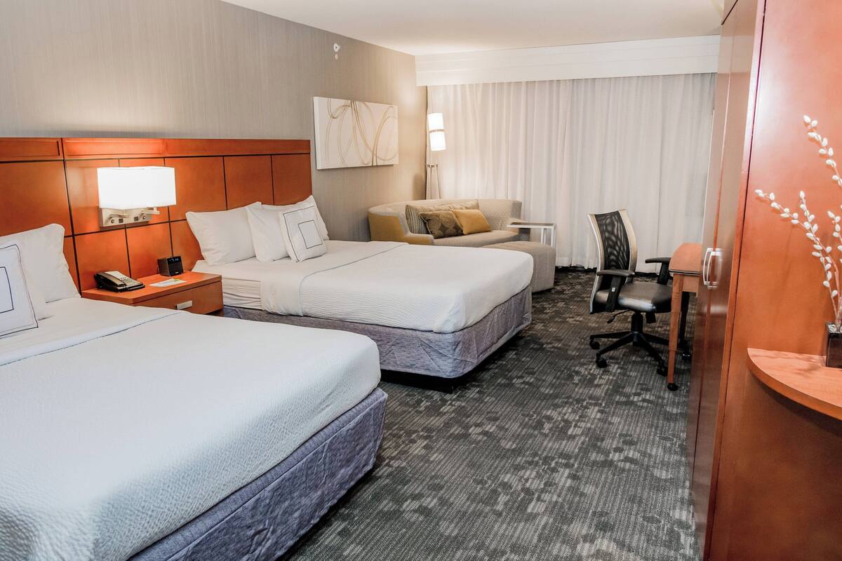 Habitación de hotel con dos camas, un escritorio, una silla y un ventanal con cortinas.
