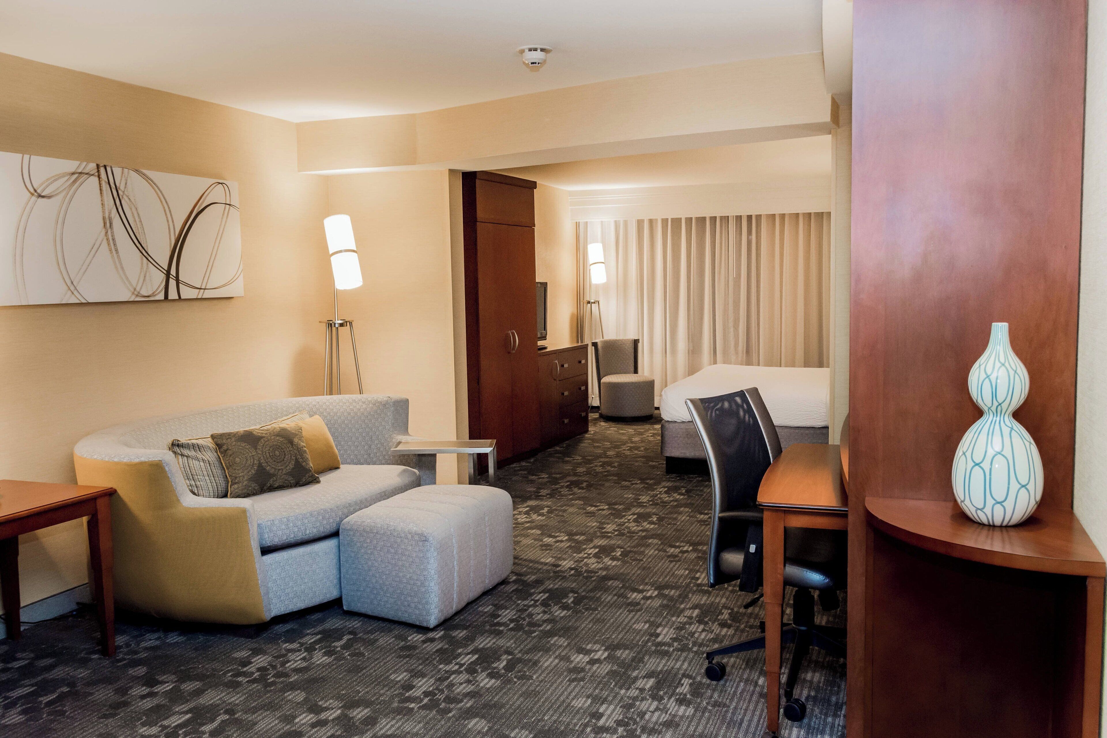 A hotel room with a sofa, ottoman, desk, chair, lamp, and a decorative vase.