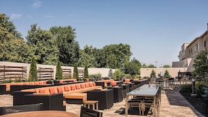 An outdoor patio with multiple seating areas, including sofas and tables, surrounded by trees and a building in the background. - Courtyard Marriott Lafayette (Lafayette)