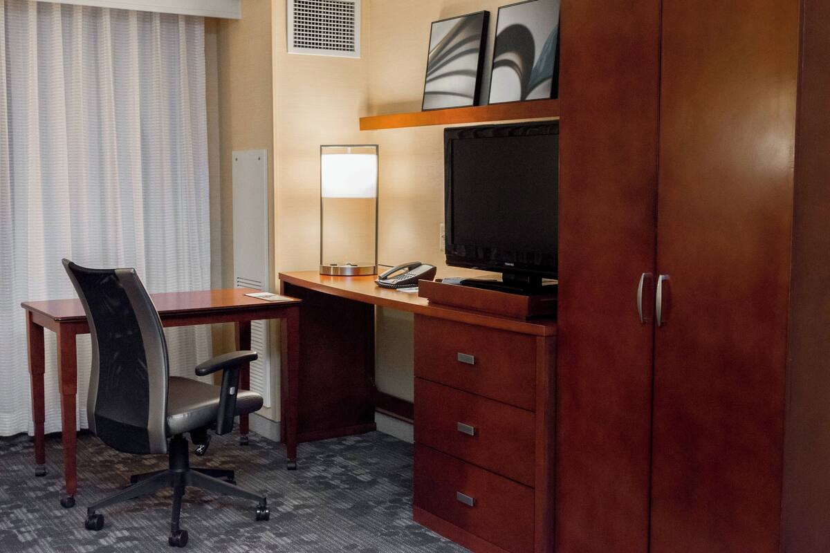Habitación de hotel con un escritorio de madera, una silla, un televisor, una lámpara y un armario.